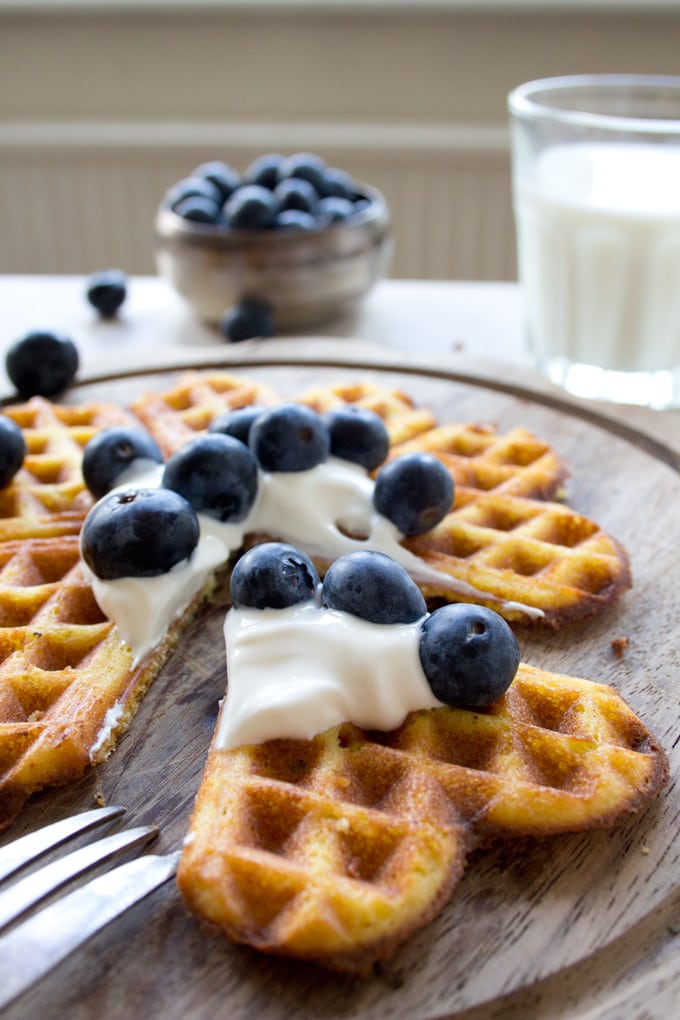 a heart shaped waffle piece being on a wooden board with blueberries and yoghurt