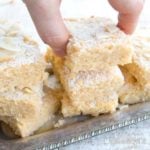 hand lifting a keto blondie from a silver tray stacked with coconut blondies