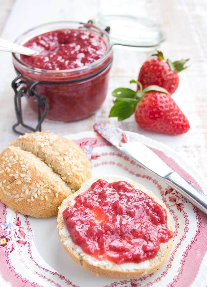 a breakfast bun spread with sugar free strawberry jam on a plate with a knife and a jar or strawberry jam