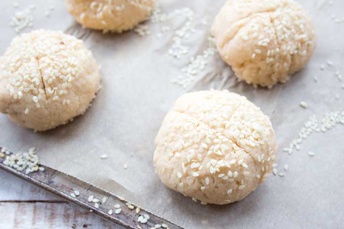 Unbaked buns topped with sesame seeds on a baking sheet lined with baking paper.