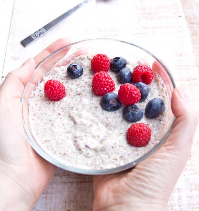 two hands holding a bowl of keto low carb porridge decorated with blueberries and raspberries