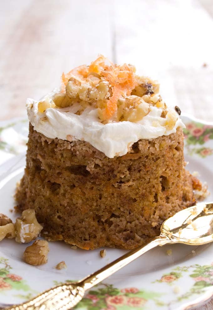 A single serve carrot cake turned out onto a plate decorated with cream cheese frosting, carrot shavings and crushed walnuts and a spoon.