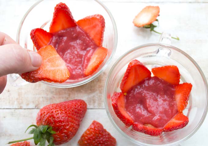 a hand adding sliced strawberries whilst assembling a strawberry shortcake trifle inside two glasses