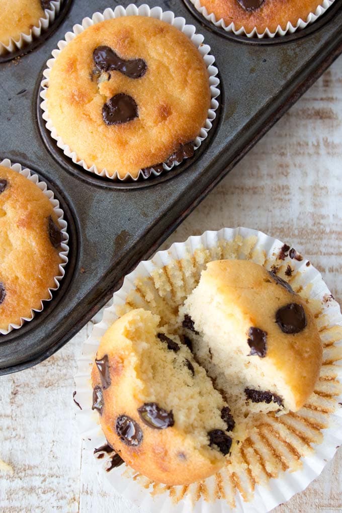 a chocolate chip muffin broken in half inside its paper casing next to a baking tray with more muffins