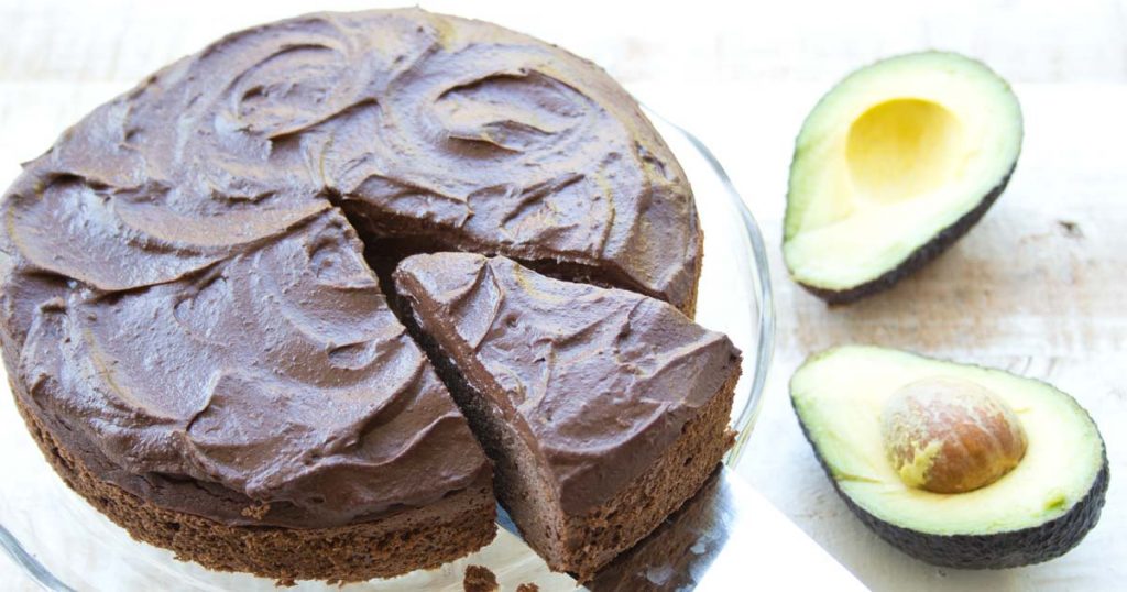 a chocolate cake with a slice being taken out by a silver cake server on a glass tray and two halves of avocado on a table