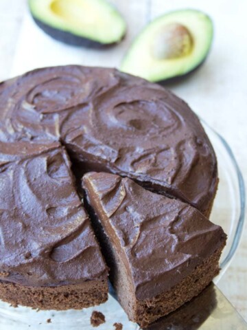 a cake server taking a slice of chocolate avocado cake with chocolate frosting from a cake on a glass tray with avocado halves in the background
