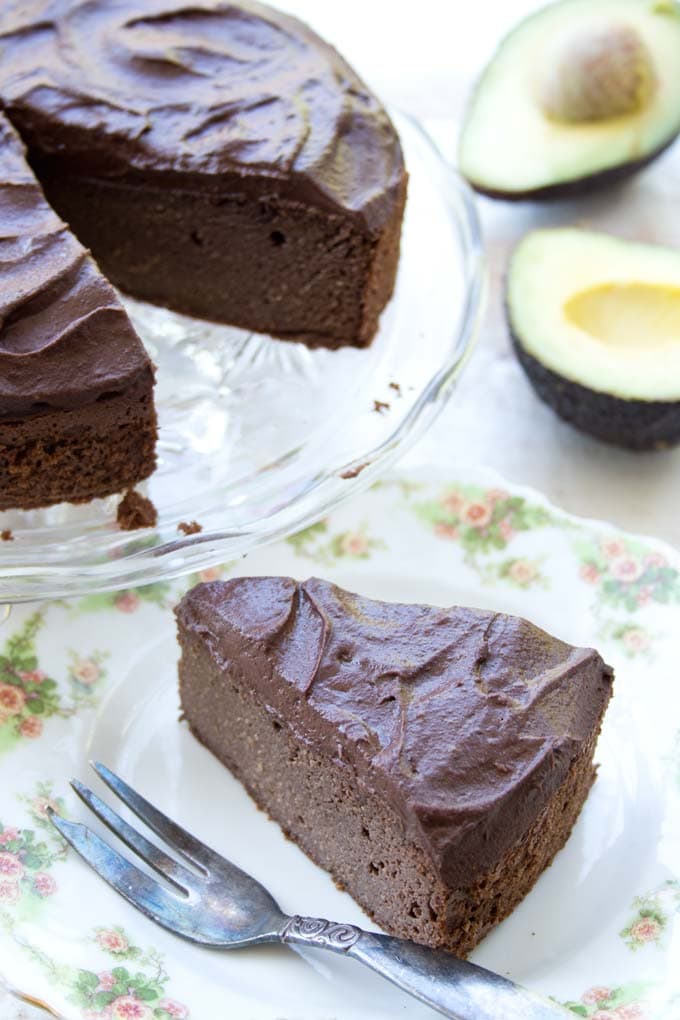 a slice of chocolate cake with chocolate frosting on a plate with a silver fork with a cake on a glass cake stand and two halves of avocado in the background