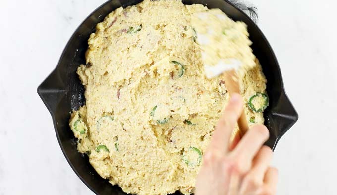 A hand spreading the batter in a black cast iron skillet with a spatula