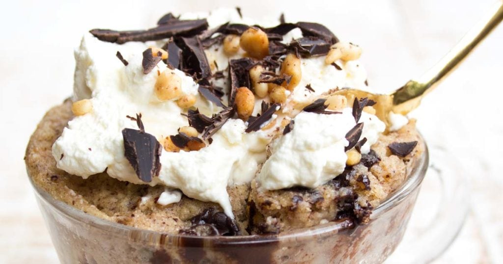 a peanut butter mug cake topped with whipped cream, peanut butter pieces and chocolate shavings and a spoon