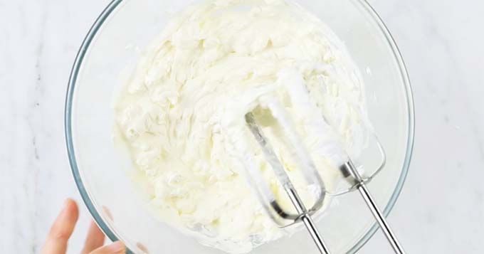 the mixing attachments of an electric hand mixer being removed from a mascarpone and cream mix in a glass bowl