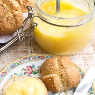a roll spread with lemon curd on a plate with a knife and a mason jar filled with lemon curd and a silver tray with more rolls in the background