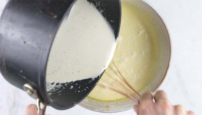 Pouring the vanilla cream mix out of the pot into a mixing bowl with the beaten egg yolks and a whisk