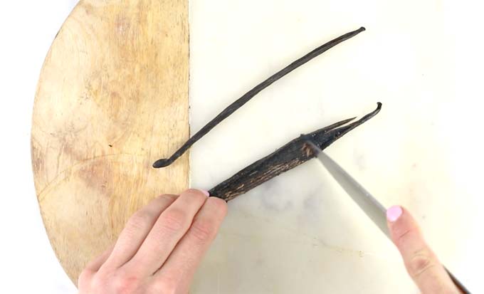 Two hands cutting a vanilla bean with a knife on a cutting board, scooping out the seeds