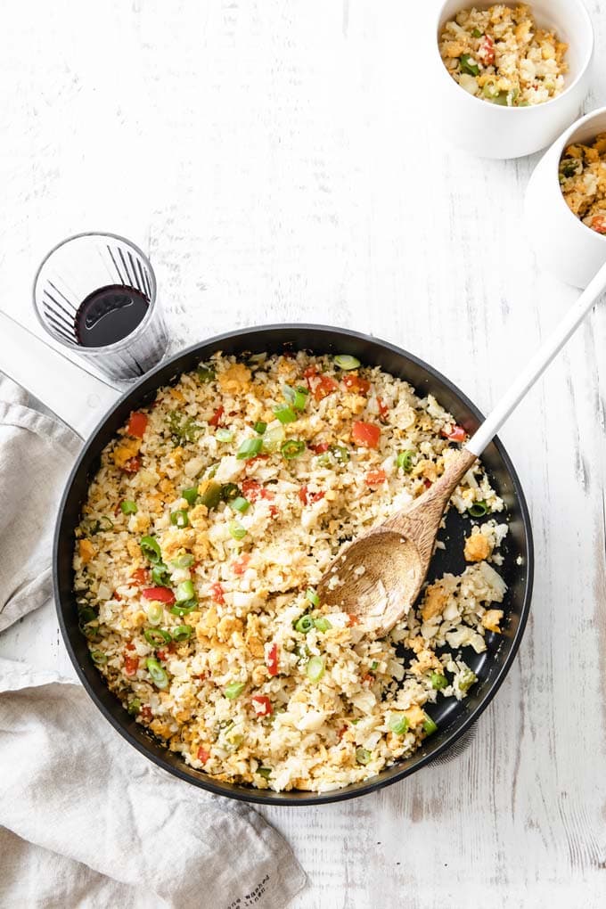 a cast iron pan with keto fried rice and a wooden spoon on a white wooden table