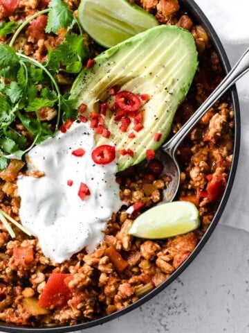 A pan with low carb chili made from turkey mince and topped with sour cream, sliced avocado, cilantro and a lime wedge