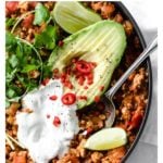 a cast iron pan with turkey chili topped with avocado, sour cream, cilantro and lime and a spoon