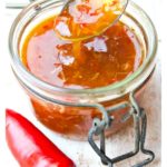 a glass jar with sugar free sweet chilli sauce and a spoon dripping with sauce with a red chilli on the side