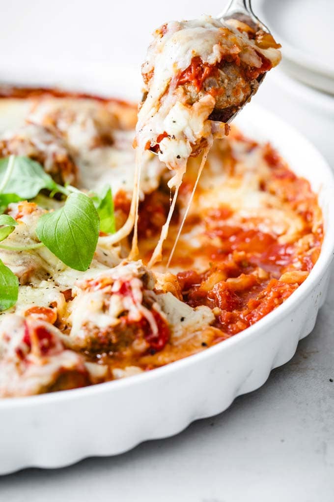 a spoon lifting a meatball covered in melted cheese from a casserole of keto meatballs with tomato sauce and cheese, decorated with green leaves