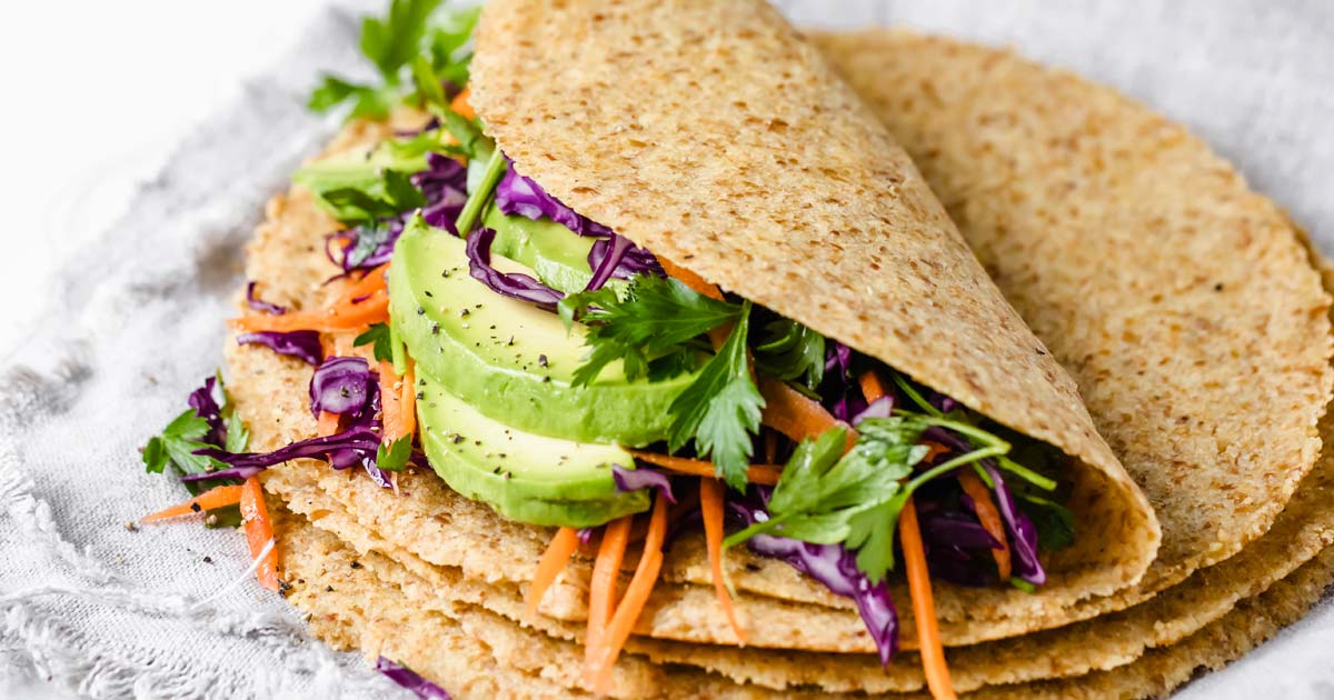 a stack of keto tortillas, the top tortilla is filled with sliced avocado, carrots, lettuce and other