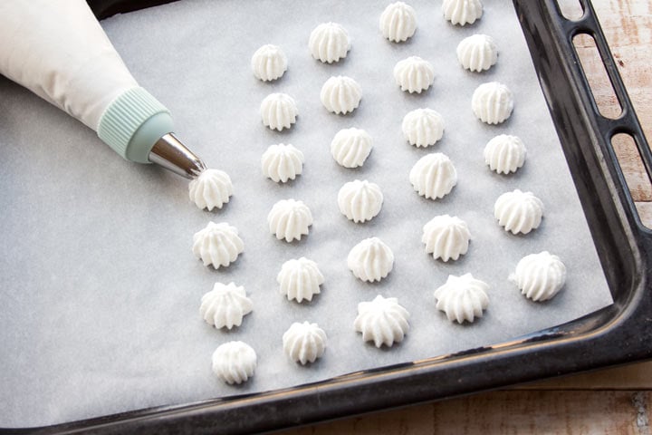 A piping bag squeezing meringue cookies onto a lined baking sheet.