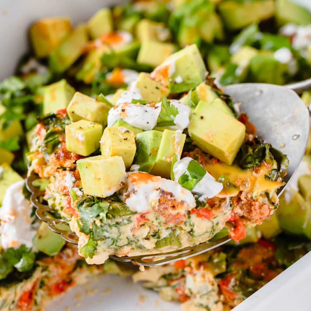 lifting a slice of chorizo breakfast casserole topped with avocado out of a casserole dish