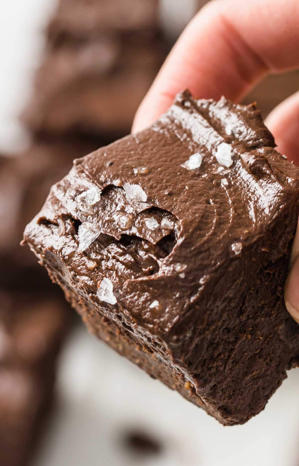 hand holding a flourless avocado brownie showing the chocolate frosting and sea salt flakes