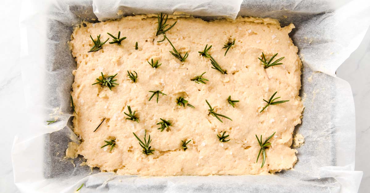 dough spread in a rectangular bread pan lined with parchment and topped with rosemary sprigs and sea salt