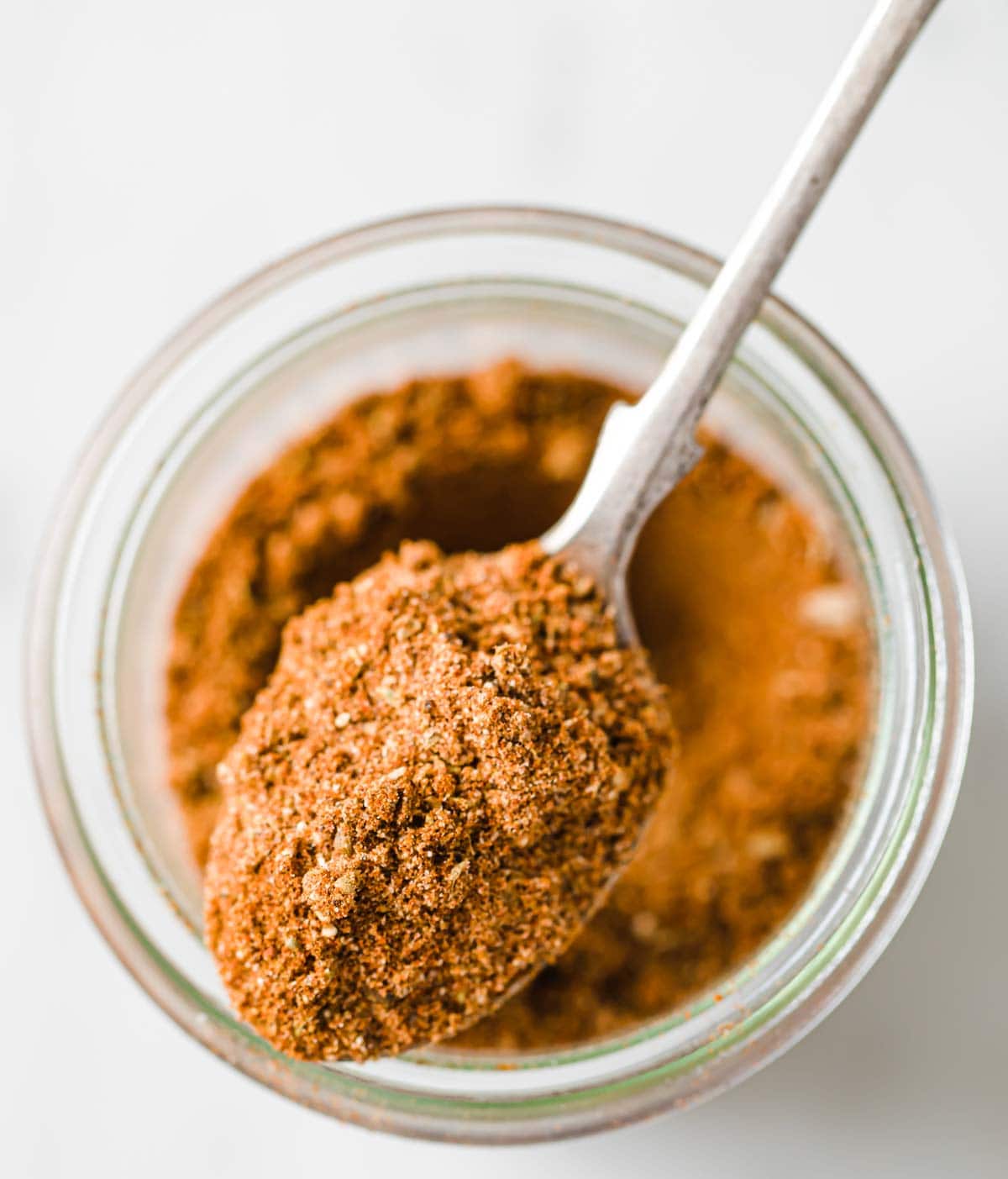 a spoon with homemade taco seasoning lying on top of a glass jar with seasoning