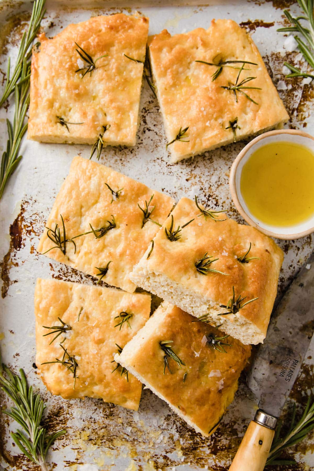 Squares of focaccia bread on a baking sheet decorated with fresh rosemary sprigs and a bowl of olive oil