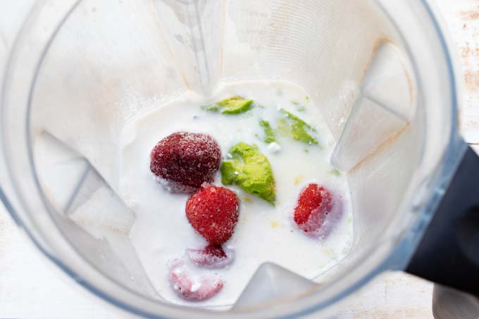 a food processor bowl with frozen strawberries, avocado, coconut cream and almond milk