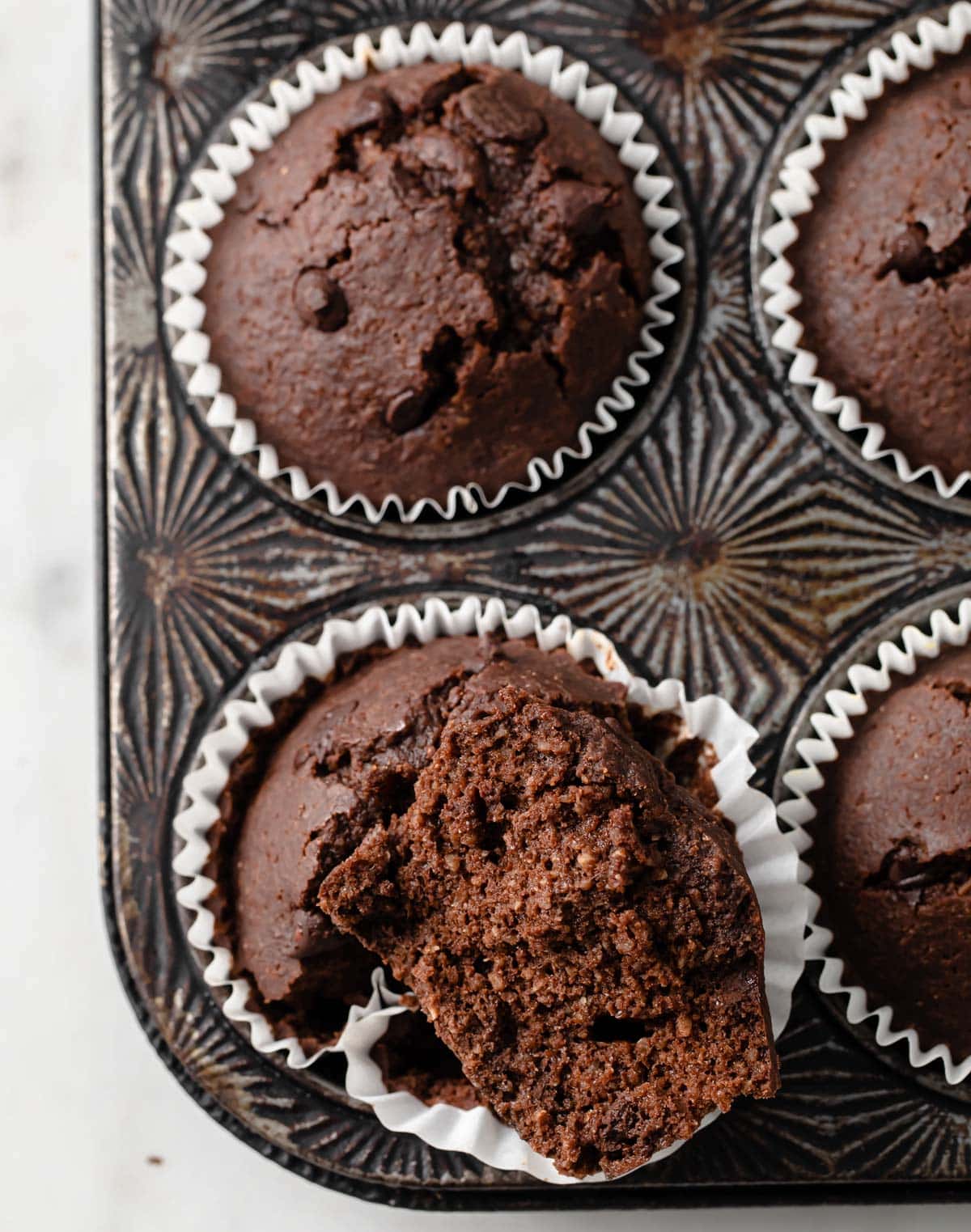 a muffin pan with almond flour chocolate muffins, one is broken in half showing the inside