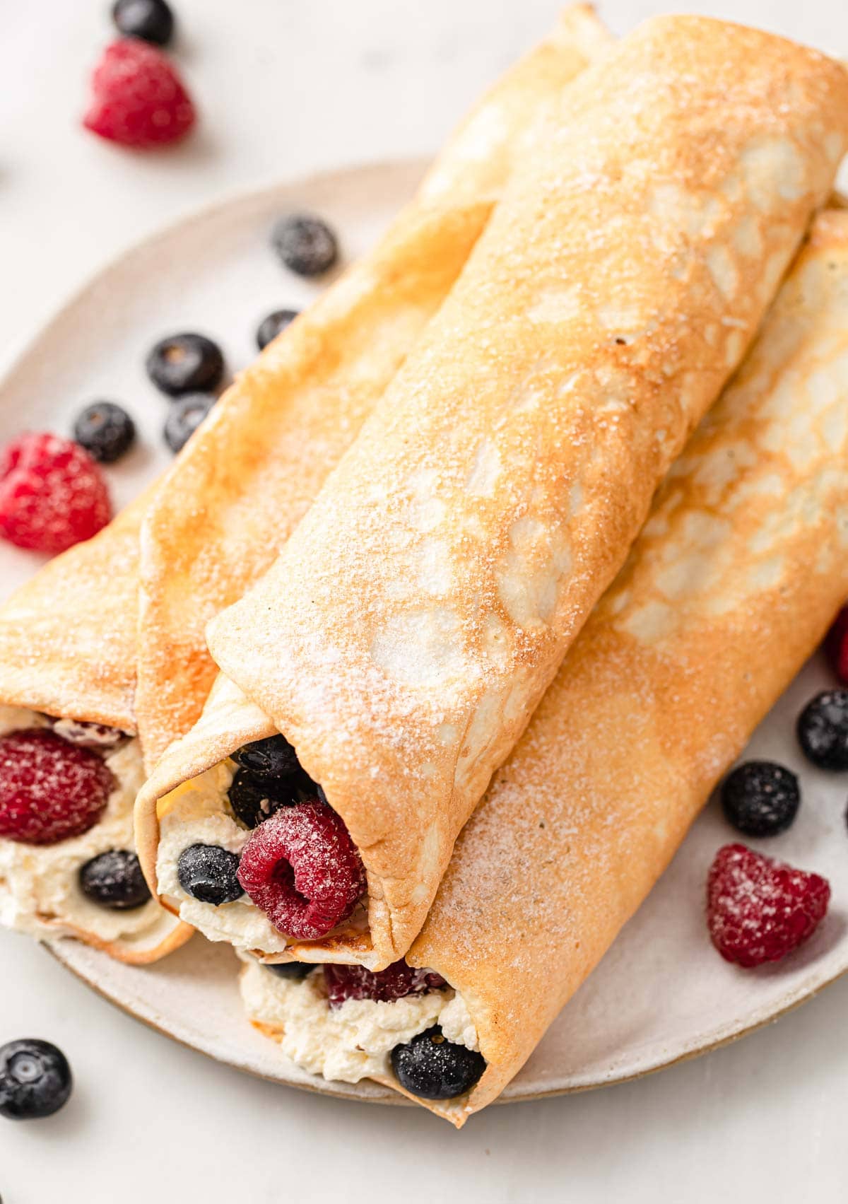 three crepes rolled up and stacked on a plate filled with cream and berries