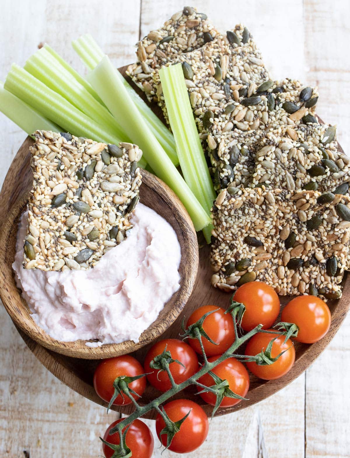 keto seed crackers on a wooden board with taramasalata, celery sticks and cherry tomatoes