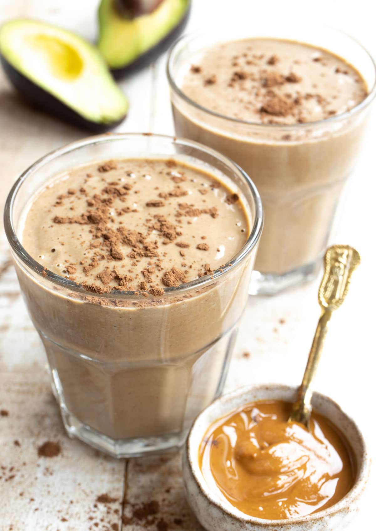 two glasses of peanut butter smoothie plus a sliced avocado and a pot with peanut butter