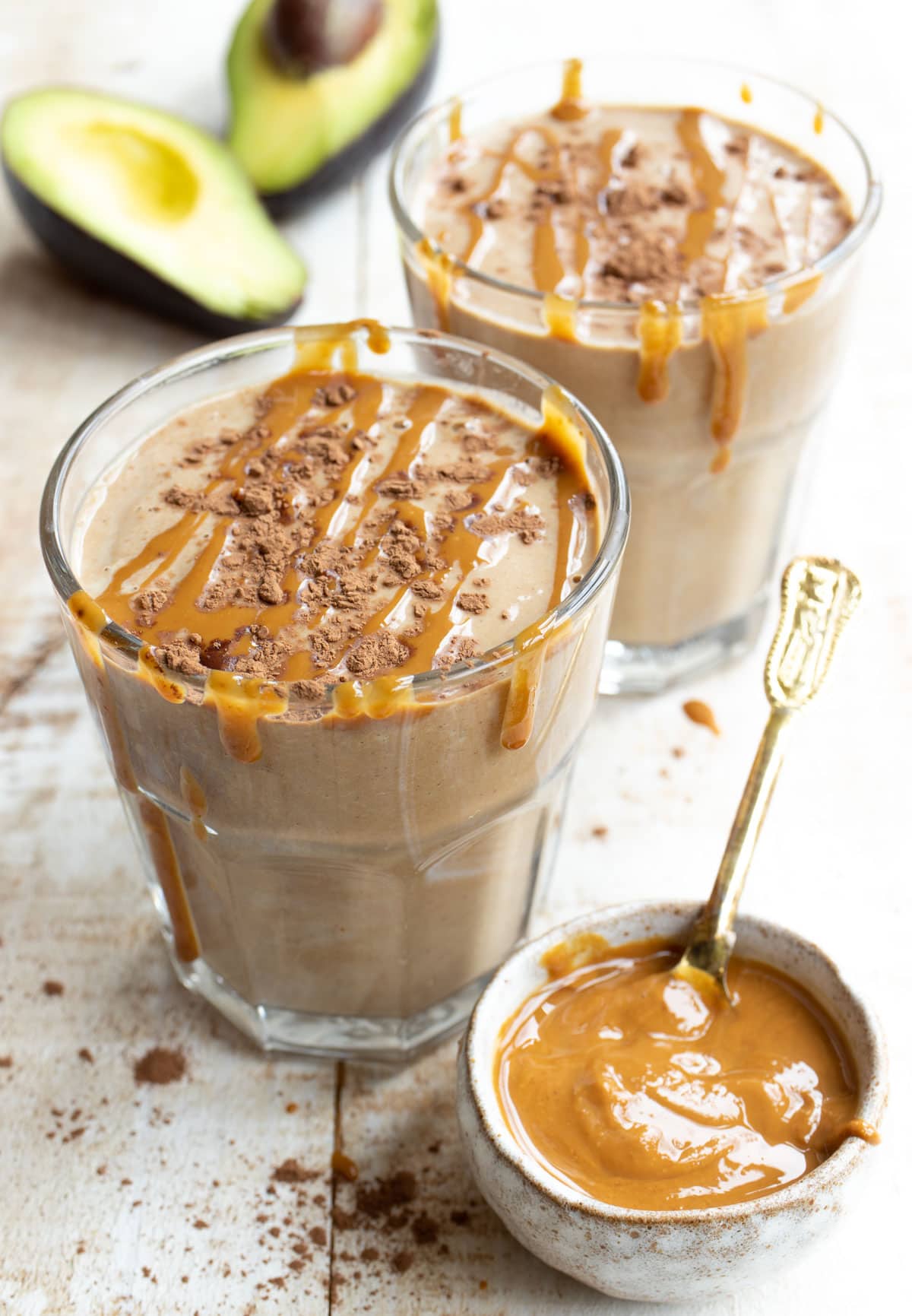 two peanut butter shakes on a table with a small pot of peanut butter and a sliced avocado