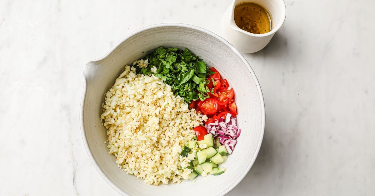 a bowl with cauliflower rice, chopped vegetables and dressing before mixing