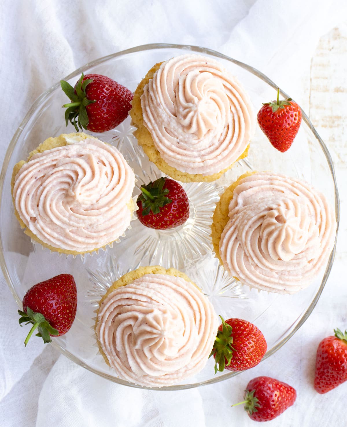 4 cupcakes with strawberry buttercream frosting on a cake stand with strawberries