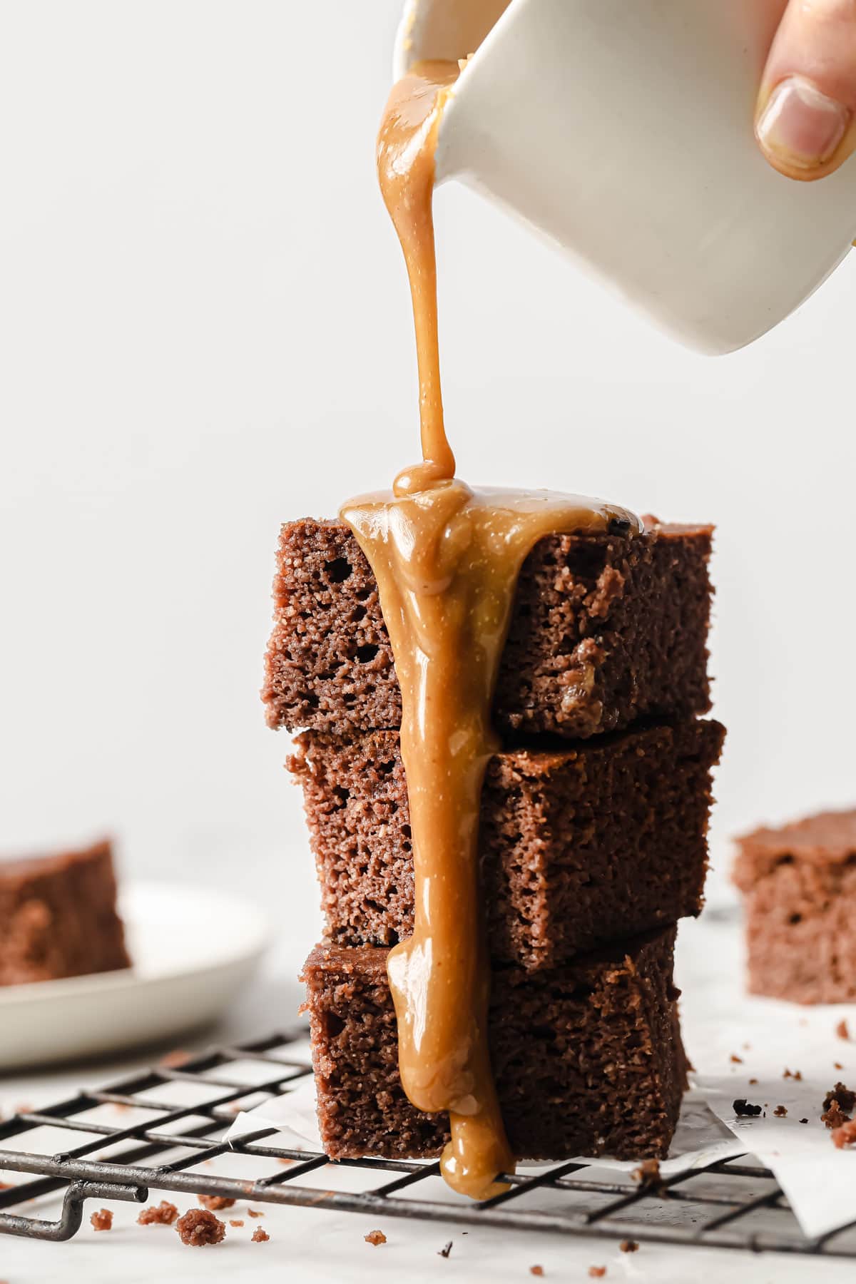 a stack of keto sticky toffee pudding squares and a hand pouring caramel sauce over it from a jug