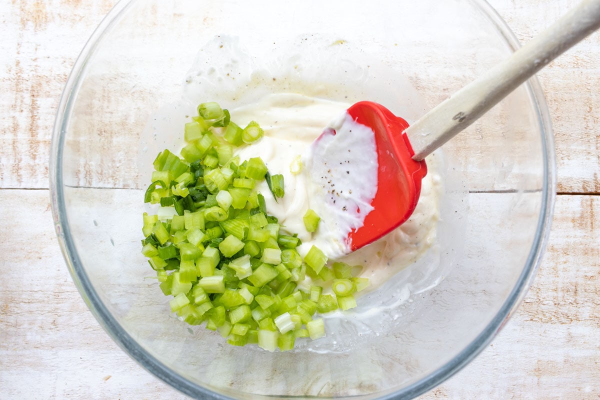 Chopped celery and spring onions in a bowl with mayonnaise and a spatula. 