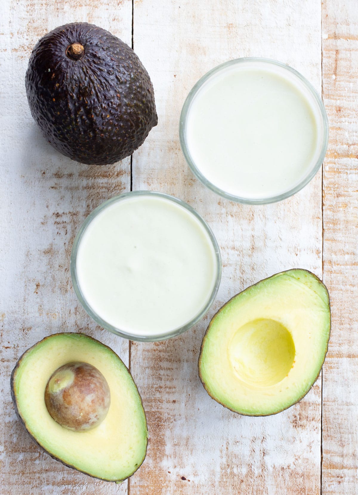 2 glasses with avocado milk on a wooden surface with halved and whole avocados arranged around.