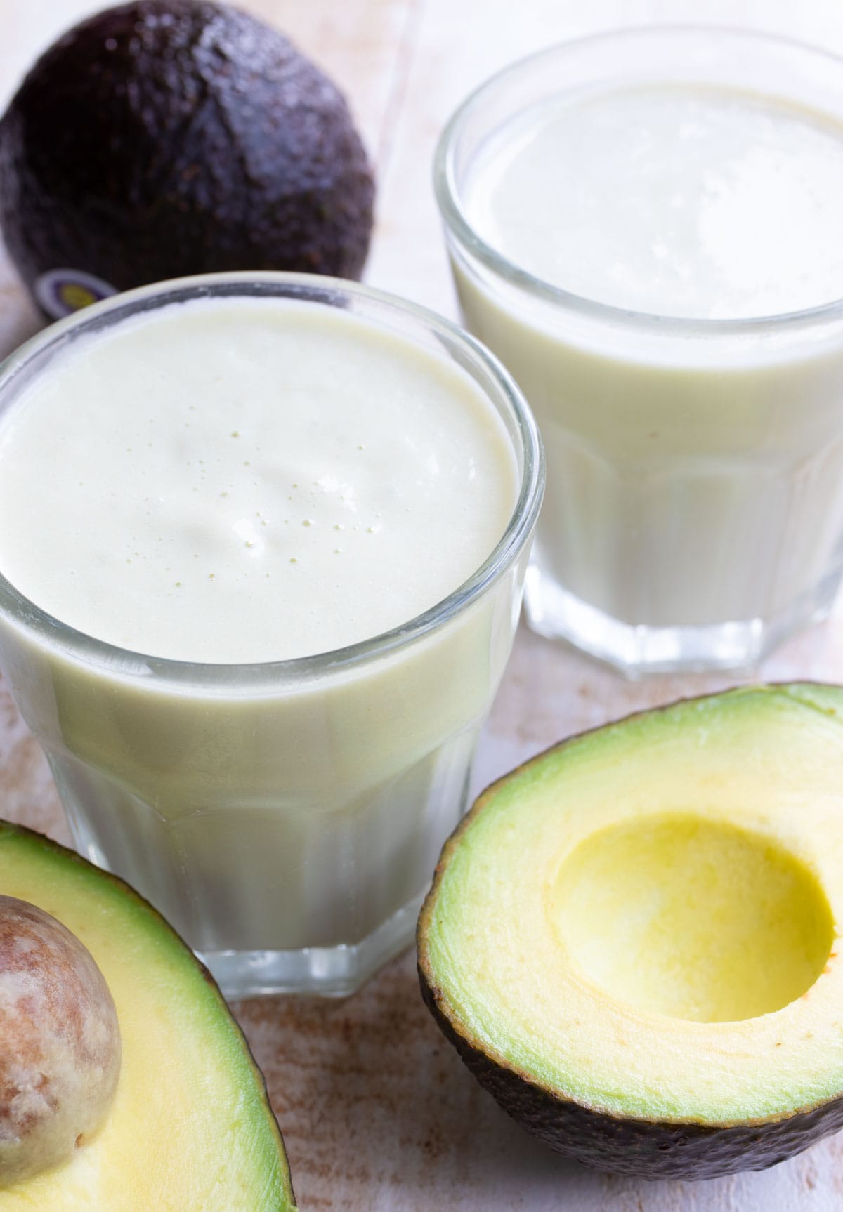 2 glasses with avocado milk and an avocado sliced in half.