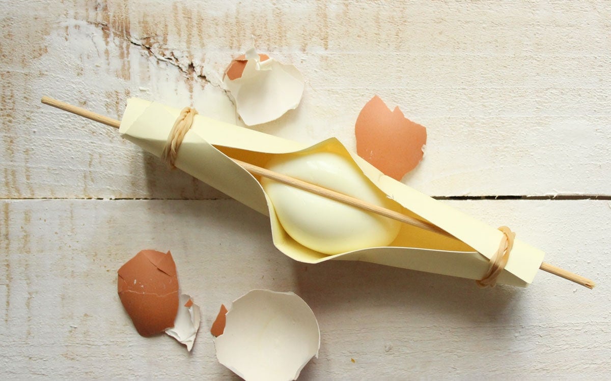 A skewer pressing on a hard boiled egg held in place by cardboard.