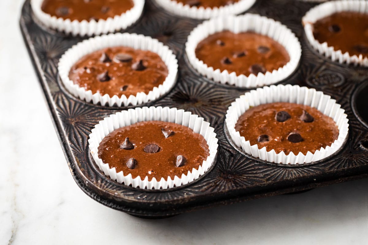 Chocolate muffin batter topped with chocolate chips filled in paper cases in a muffin tin.