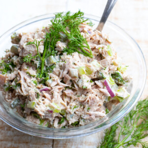 Tuna salad with chopped red onion, celery and pickles topped with fresh dill sprigs in a glass bowl.