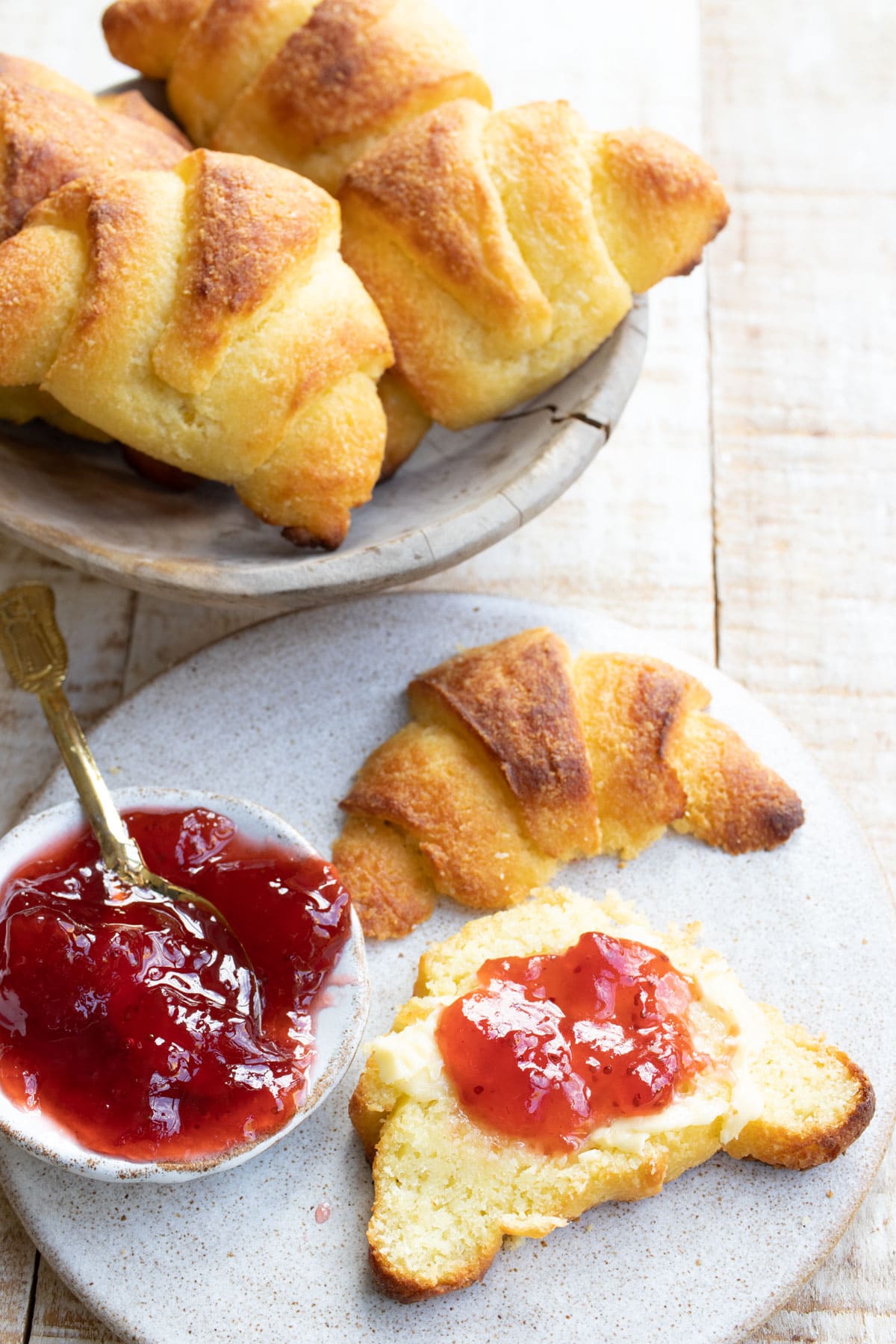 A croissant sliced open and topped with butter and strawberry jam, plus a bowl with croissants.