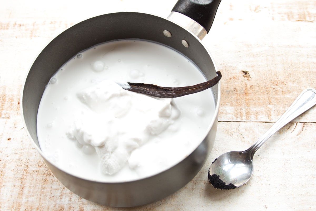 Coconut cream and vanilla pod in a saucepan and a spoon with vanilla seeds.