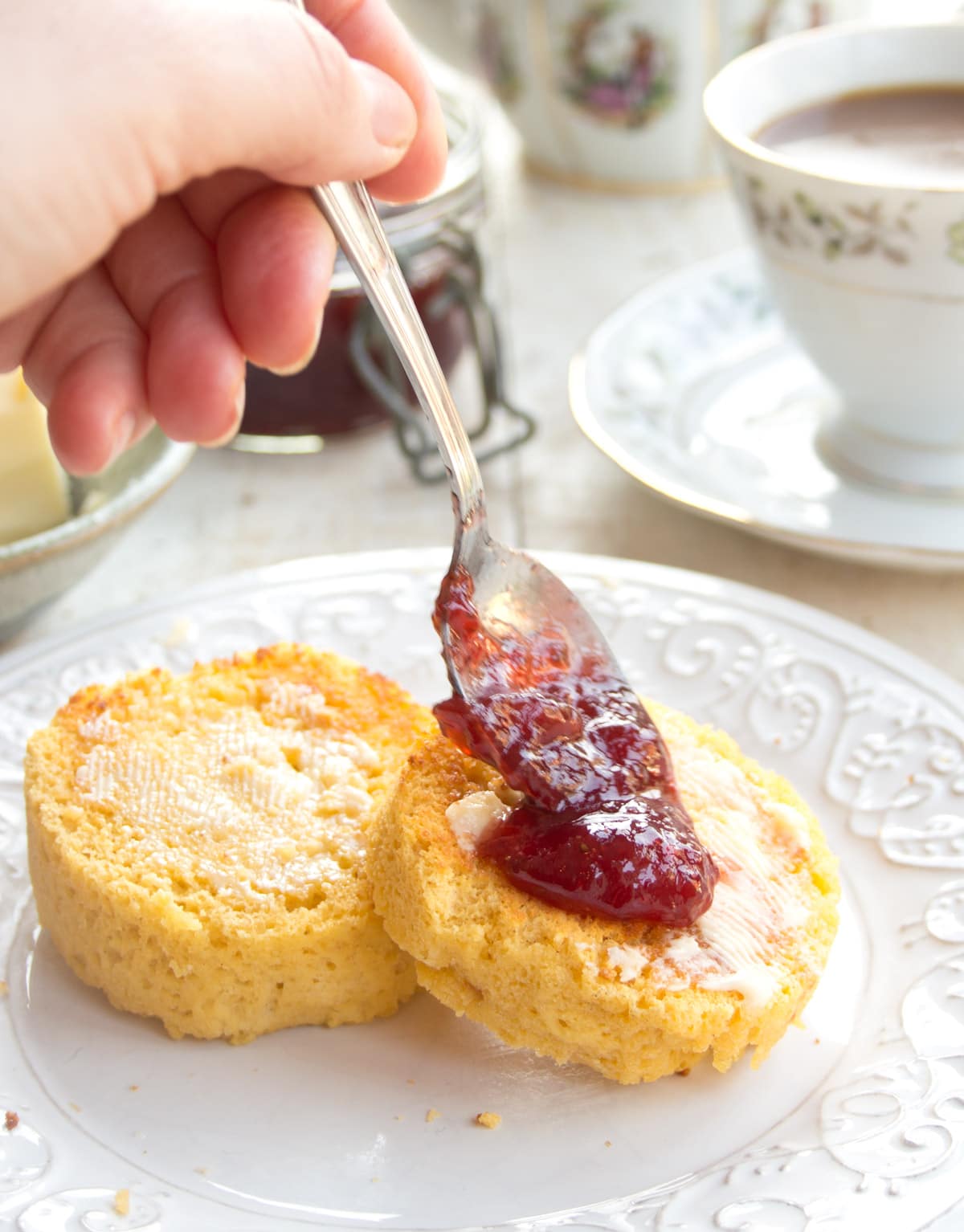 An English muffin toasted, buttered and being topped with a spoonful of strawberry jam.