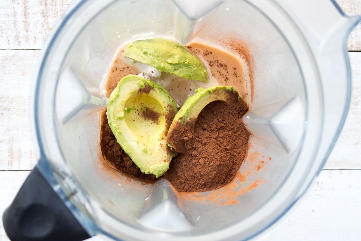 Avocado halves, cocoa powder and other ingredients in a blender jug.