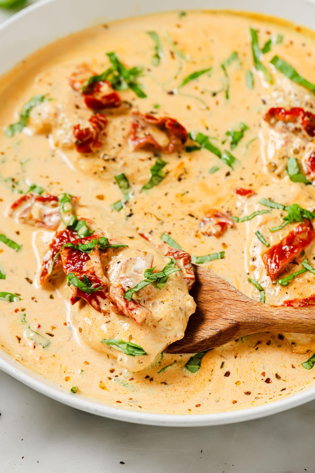 Chicken fillets in a creamy sauce with sun-dried tomatoes in a pan and a wooden spatula lifting a fillet.