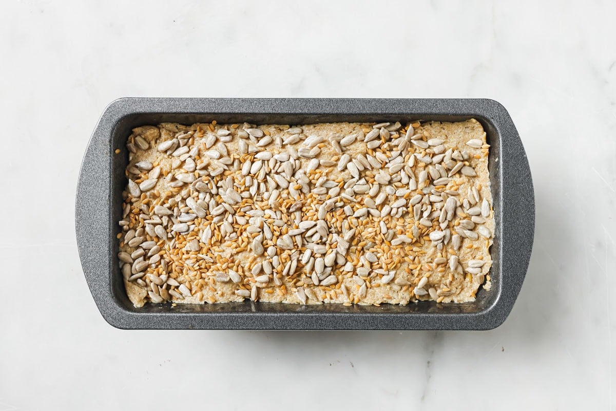 Dough in a rectangular bread pan topped with sundlower seeds and golden flaxseed.